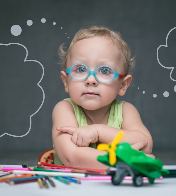 Child with glasses sitting at classroom desk with chalkboard behind.